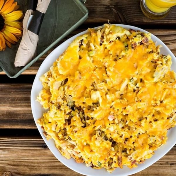 A large plate of cheesy hash browns with a sunflower and orange juice on a wooden table.