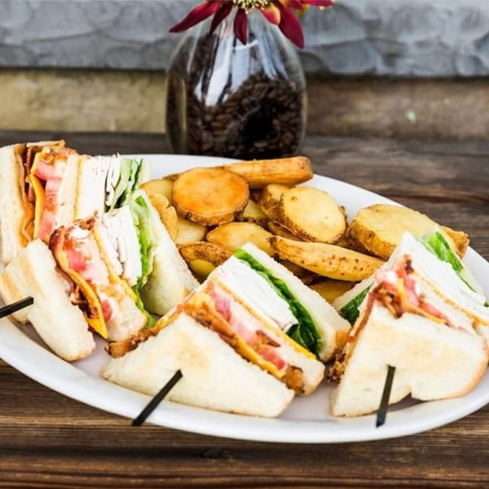 A white plate holds several club sandwich halves with bacon, lettuce, and tomato, alongside potato wedges.
