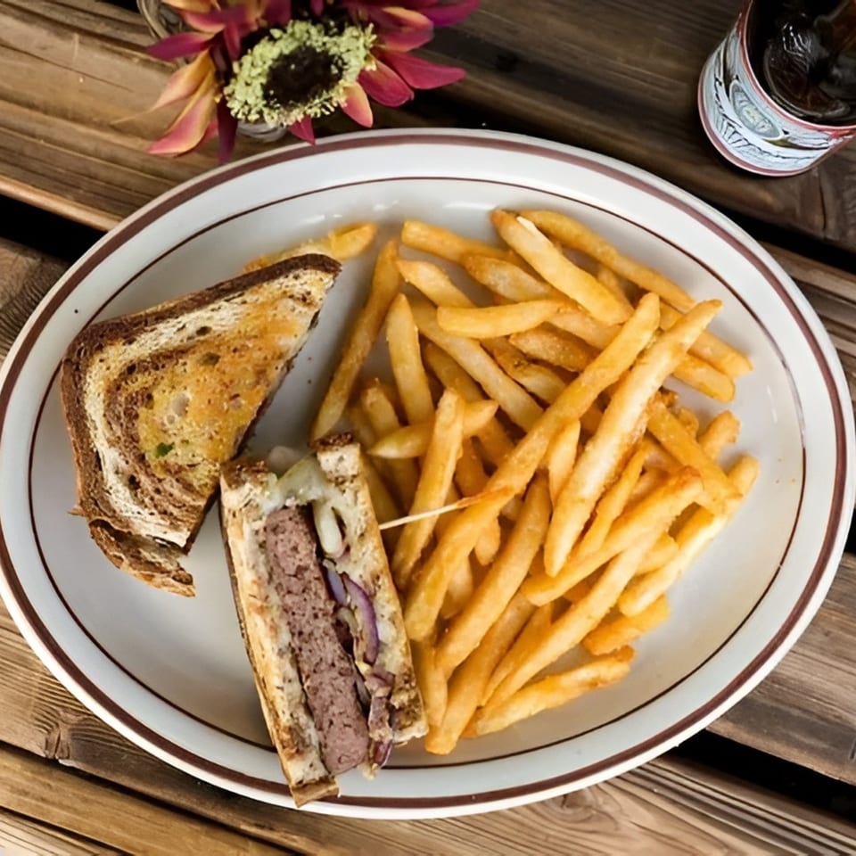 Patty melt on marbled rye with red onion and fries, served on a white plate on a wooden table.